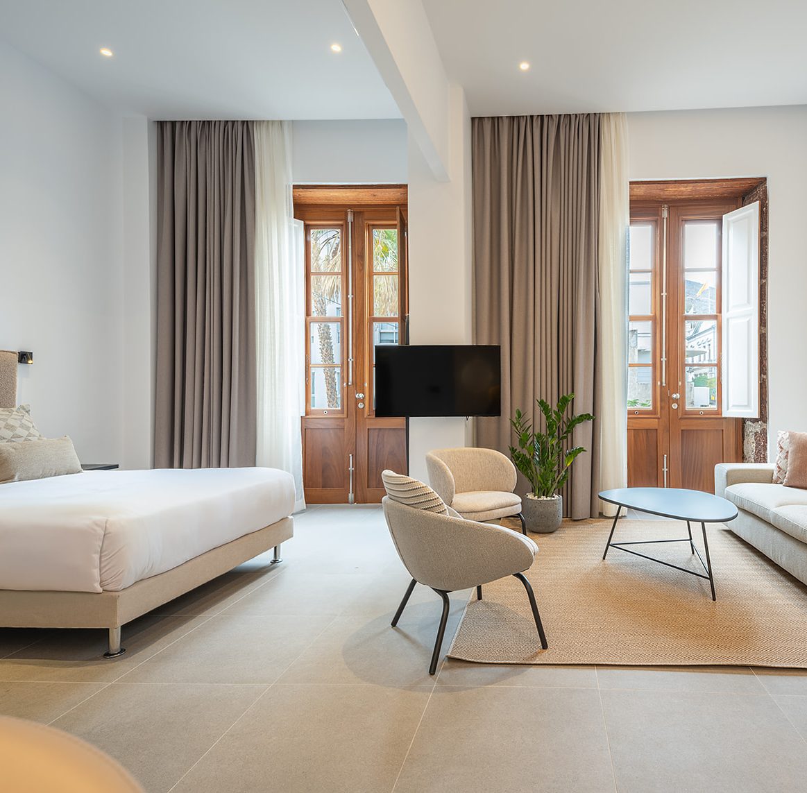 An open-plan suite with no partition between the bedroom and the living-dining room. The bedroom is on the left, with a swivelling TV mounted on a column and a sofa to the right of the suite. Two floor-to-ceiling windows featuring original 19th-century timber frames let in plenty of light, illuminating the entire room.