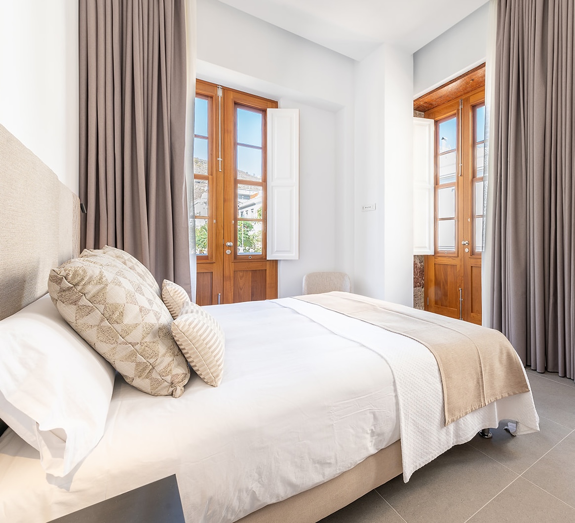 A bedroom with a king-size bed and two large floor-to-ceiling windows featuring original 19th-century timber, one at the foot of the bed and the other to its left. Grey blackout curtains reaching from floor to ceiling. A chair in the corner of the bedroom, a beige headboard, and pillows and cushions on the bed.