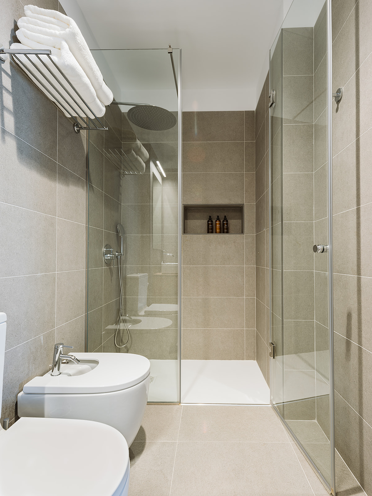 Baño con lavamanos, bidé y retrete, con ducha de mampara con una puerta abatible y hornacina con gel, champú y acondicionador de cortesía. Barra sobre inodoro con toallas blancas de baño.