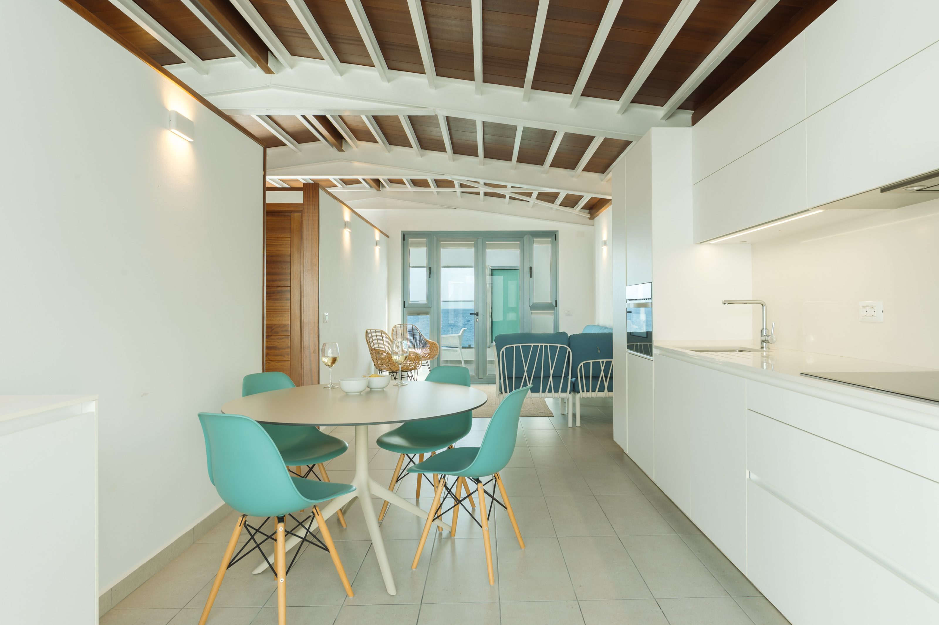 Zona común de apartamento en distribución de espacio abierto continuo, comenzando por la cocina a la derecha. A su izquierda frente a la cocina, una mesa redonda de comedor con cuatro sillas azules. Tras la cocina y el comedor, un espacio de sofá a la derecha y dos butacas individuales de mimbre a la izquierda. El espacio de sofá da paso a un ventanal con puerta corredera, todo de cristalera, que permite ver una terraza al fondo con vistas al mar.