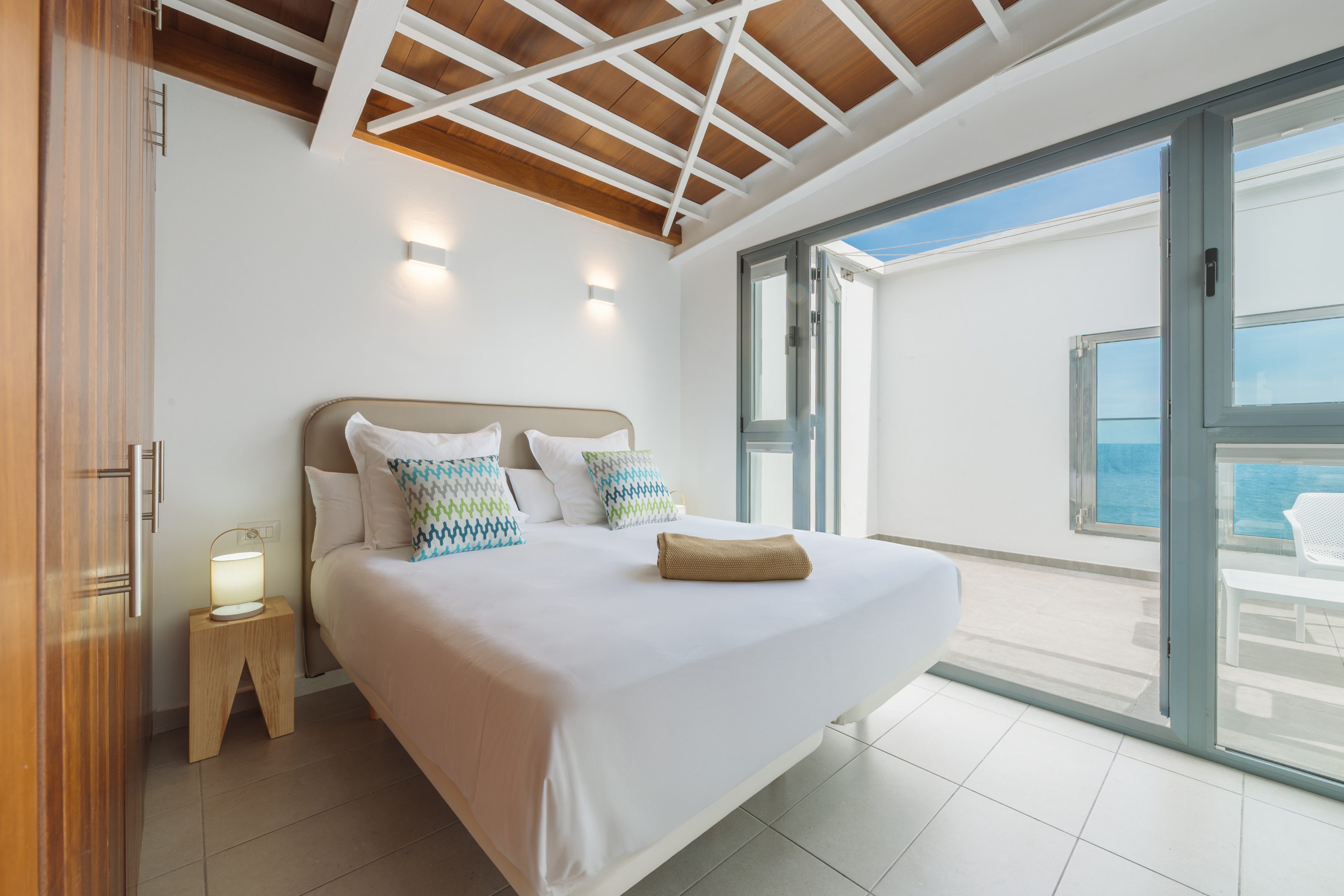 Dormitorio con cama King Sice, mesilla de noche con lampara a la derecha de la cama y armario empotrado a lo largo de la pared lateral. A la izquierda de la cama, una pared de cristaleras de suelo a techo con puerta corredera que da acceso a una terraza con su propio ventanal con vista mar.
