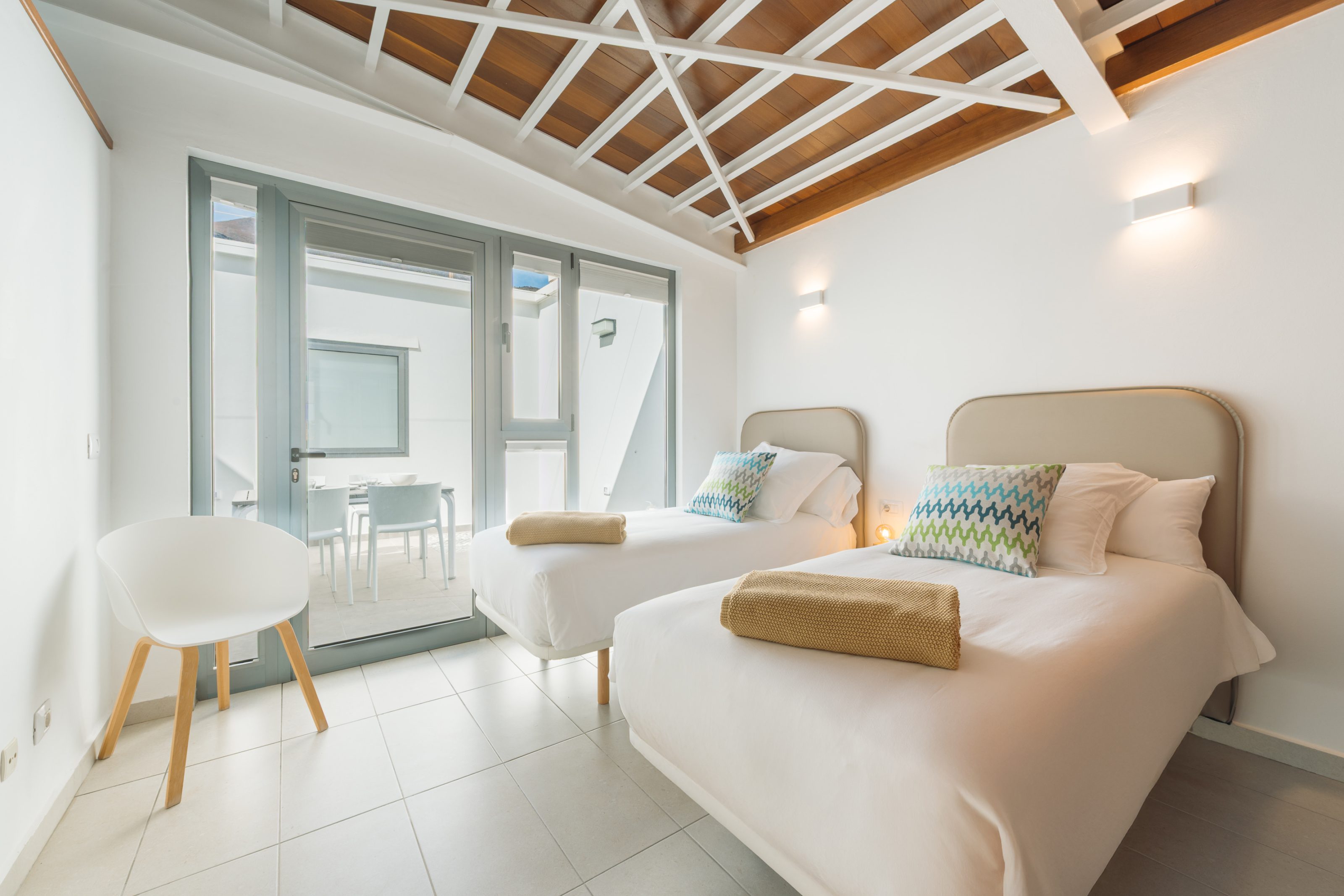 Dormitorio con dos camas individuales de 90cm, una butaca en esquina y un ventanal de suelo a techo en la pared de fondo que da acceso a una terraza interior con mesa de comedor.