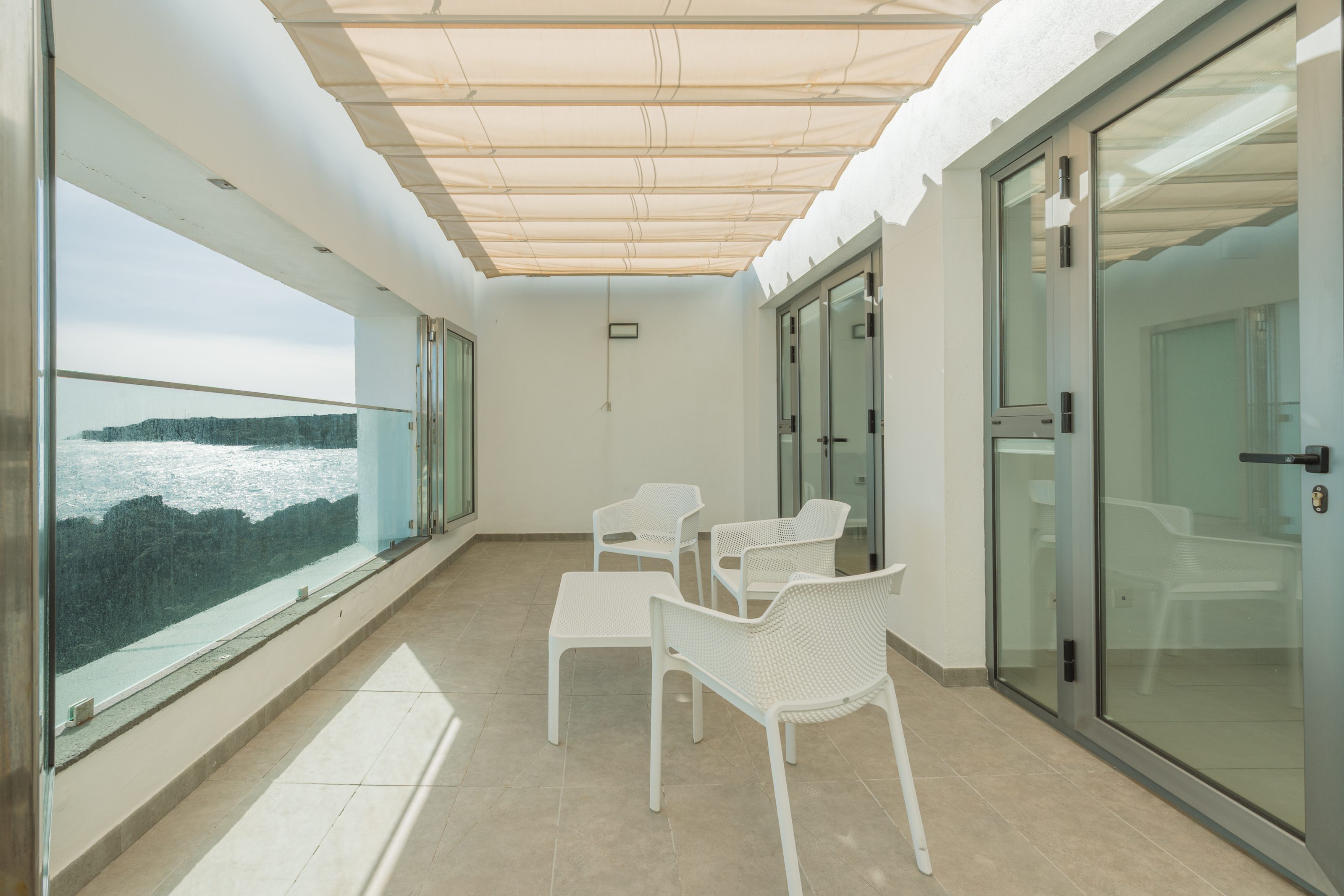 Terraza exterior con parasol abatible de techo. A la izquierda, un ventanal abierto de vista panorámica a pie del rompeolas, mirando el Océano Atlántico. A la derecha, puertas correderas de cristaleras de acceso a la vivienda. En el centro de la terraza hay tres sillas y una mesita de café blancas de exterior.