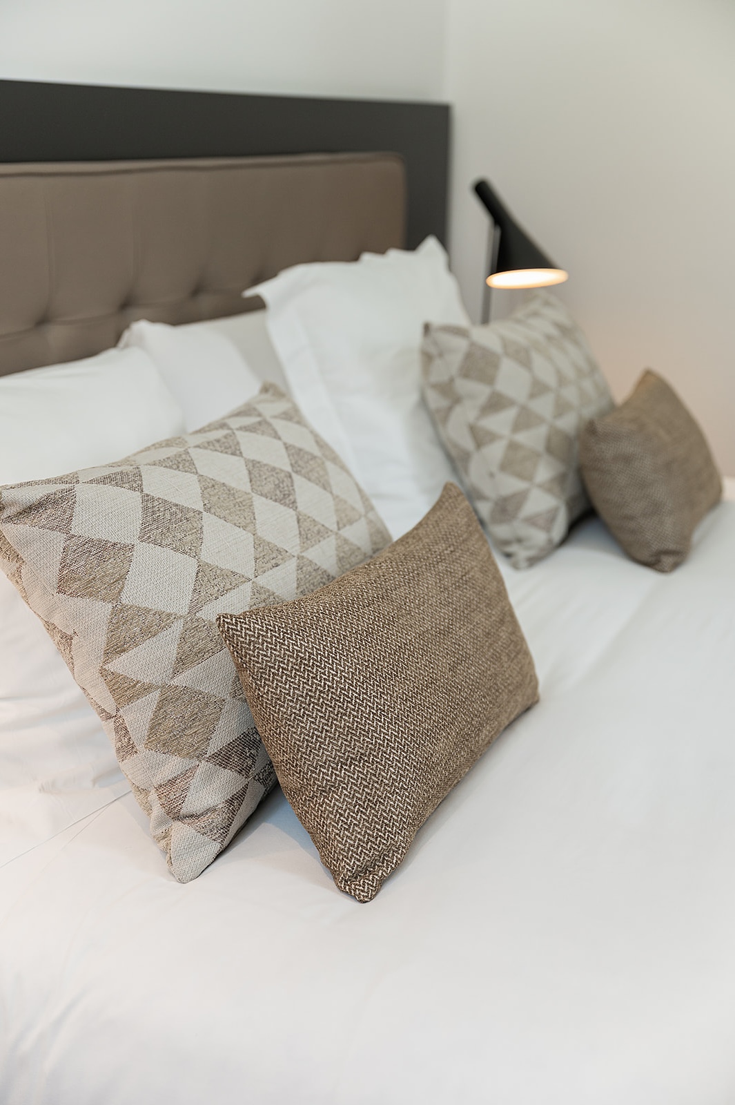 King-size bed in Suite 4 at The Park Suites, with pillows, duvet covers and decorative cushions for two people. Beige headboard and white sheets. Black bedside lamp.